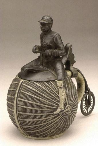 Beer Stein, ca. 1890<br>
Pewter rider/lid and rear wheel<br>
Stoneware container<br>
Germany<br>